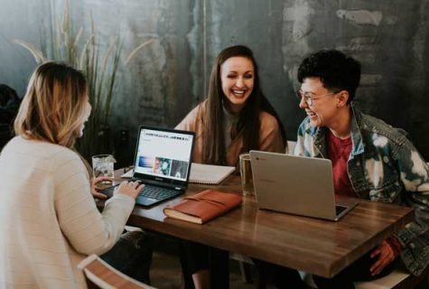 Three people sit at table working on laptops together while laughing.